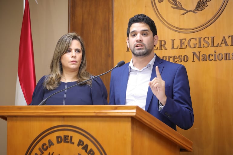 Una mujer de mediana edad vestida con una camisa azul de mangas abullonadas. A su lado un hombre joven con barba, camisa blanca, saco azul, que levanta un dedo índice; ambos detrás de un atril con el escudo de Paraguay. A la izquierda una bandera paraguaya. De fondo un escudo del Poder Legislativo en la pared revestida con madera.