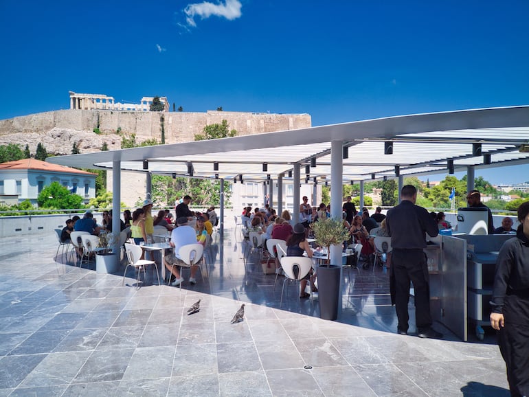 Atenas, Grecia: restaurante en el Museo de la Acrópolis con terraza abierta y vista al Partenón, lleno de clientes en un día soleado.