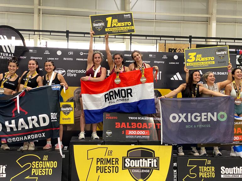 Las paraguayas Leticia Robledo, Patricia Buena y Jessica Chávez, con las medallas y el trofeo correspondiente al primer puesto, así como la bandera paraguaya en el podio del SouthFitnes Challenge 2024.