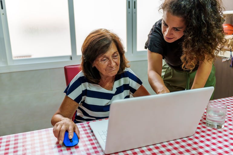 Mujer de mediana edad enseña a un adulto mayor a usar una computadora portátil, cerrando la brecha digital generacional.