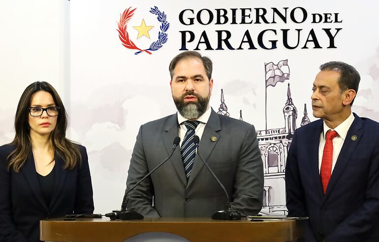 Una mujer joven, con traje negro, pelo suelto negro y anteojos, un hombre con traje y barba y a su lado otro hombre que lo mira de reojo con traje y corbata roja. De fondo se ve una silueta dibujada del palacio de gobierno y se lee la frase Gobierno del Paraguay.