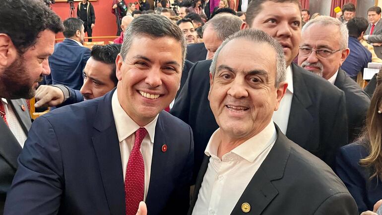 Un hombre de mediana edad con traje y corbata roja, sonriendo, junto a otro hombre adulto de saco y camisa, rodeados por otros hombres en un acto público.