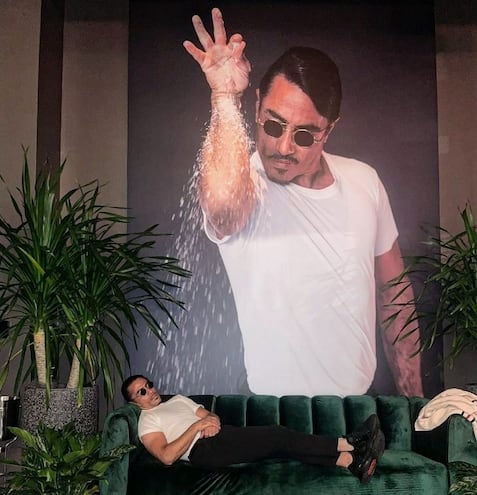 El chef turco Salt Bae posa recostado en un sillón junto a un retrato gigante de él mismo que lo muestra en su característica pose al salar la carne.