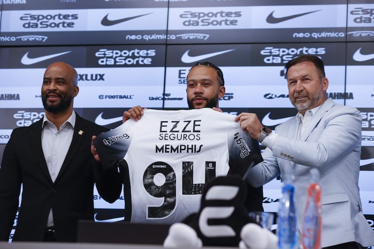 El futbolista neerlandés Memphis Depay (c), sostiene la camisa del Corinthians durante su presentación como nuevo jugador del club acompañado del presidente del Corinthians Augusto Melo (d) y el manager Fabio Soldado (i), durante una rueda de prensa este jueves en el estadio Neo Química Arena, sede del club Corinthians en Sao Paulo (Brasil). Depay fue recibido como una estrella en el aeropuerto internacional de Guarulhos, en São Paulo, por la afición del Corinthians, en un momento en que el equipo lucha por no descender a segunda división.