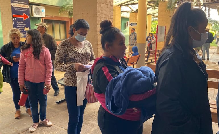 Larga fila de pacientes esperando por turnos de consultorio externo. En la fila se puede observar a una mujer con un bebe en brazos.