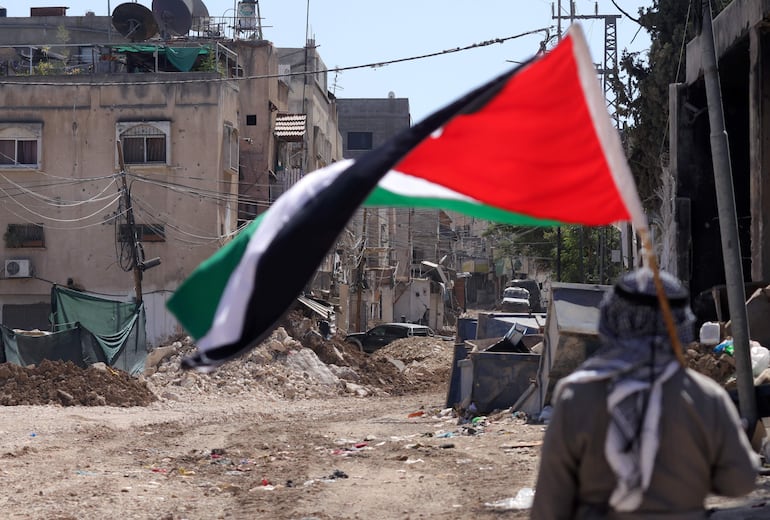 Un hombre sostiene una bandera palestina mientras soldados israelíes realizan una operación en el campo de refugiados de Tulkarem, cerca de la ciudad cisjordana de Tulkarem, este miércoles. Al menos dos palestinos murieron el 10 de septiembre durante las operaciones militares israelíes en Tulkarem y su campo de refugiados, según el Ministerio de Salud palestino. EFE/ Alaa Badarneh