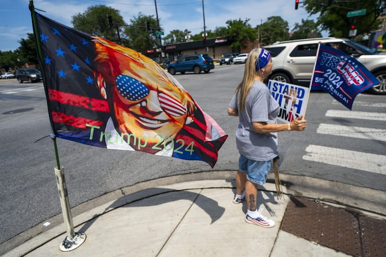 Una bandera de Trump ondea mientras una partidaria del ex presidente estadounidense sostiene un cartel. El expresidente Donald Trump, víctima ayer de un atentado fallido durante un mitin político en Butler (Pensilvania), pasó el domingo recluido en el club de golf de Bedminister, en Nueva Jersey, de su propiedad.