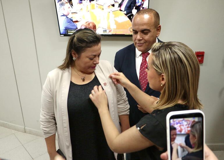 La senadora Lizarella Valiente (ANR) le coloca el pin del partido Colorado en la solapa a su colega Zenaida Delgado (ex Cruzada Nacional), bajo la mira del senador Basilio "Bachi" Núñez.