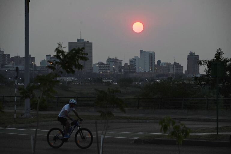 Un joven pedalea su bicicleta por la Costanera de Asunción. De fondo se ve la silueta de los edificios.