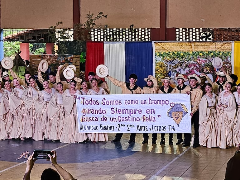 Estudiantes presentan una emotiva danza tradicional, levantando un cartel con la frase: "Todos somos como un trompo girando siempre en busca de un destino feliz".