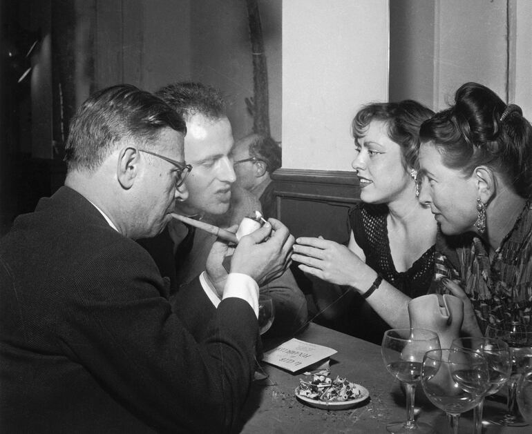 De izquierda a derecha: Jean-Paul Sartre enciende su pipa, Boris Vian habla, Michelle Vian hace ademán de acotar algo y Simone de Beauvoir escucha. Café de Flore, París, 1949.