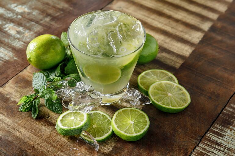 Vaso con hielo, limón y capiriña, rodeado de limones enteros, en rodajas, hielo y hojas de menta.