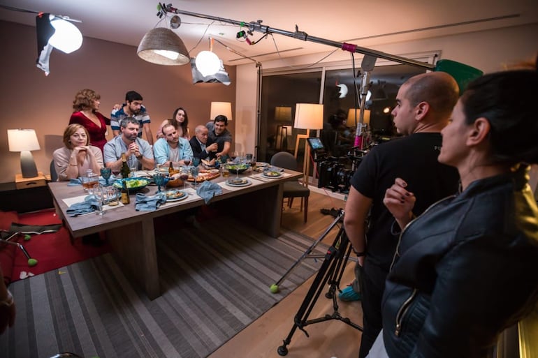 El elenco de “Junto con nuestros padres y amigos” sentado a la mesa, bajo la atenta mirada de Armando Aquino y Sandra Flecha, en rodaje.