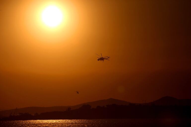 Un sol abrasador tiñe de naranja el cielo mientras helicópteros cargan agua para combatir un incendio forestal. (EFE)