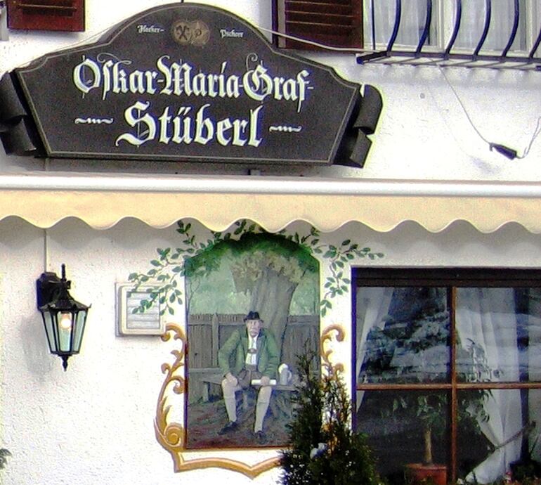 Detalle de la fachada de la "Oskar Maria Graf Stüberl", con un retrato de Oskar Maria Graf vestido con traje tradicional bávaro junto a la ventana (Foto: Robert Purdy, 2024).