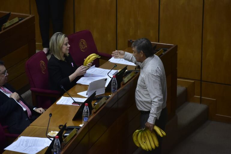 Una mujer sentada en su silla detrás de un escritorio recibe bananas de manos de un hombre adulto que tiene un cacho de bananas en la mano izquierda.