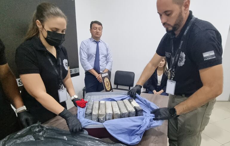 Eliana Cardozo, observa desde el fondo cuando agentes de la Senad verifican su maleta ante la atenta mirada del fiscal Osmar Segovia.