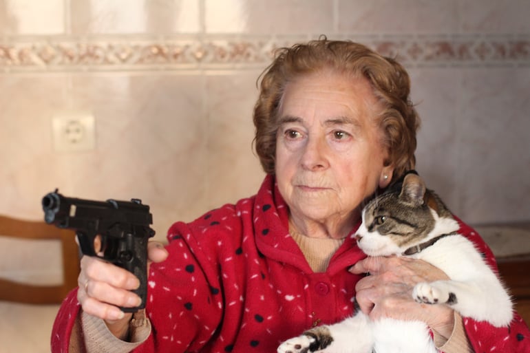 Una anciana apunta con una pistola mientras sostiene en brazos a un gato.