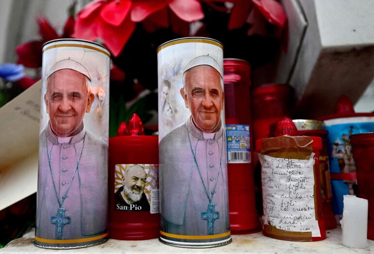 Velas con retratos del Papa Francisco y del Padre Pío se encuentran debajo de una estatua del Papa Juan Pablo II afuera del hospital Gemelli donde el Papa Francisco está hospitalizado para pruebas y tratamiento por bronquitis en Roma, el 19 de febrero de 2025. El Papa Francisco, a quien le han diagnosticado neumonía en ambos pulmones, pasó una "noche tranquila", dijo el Vaticano, en medio de crecientes preocupaciones sobre la condición del hombre de 88 años. 