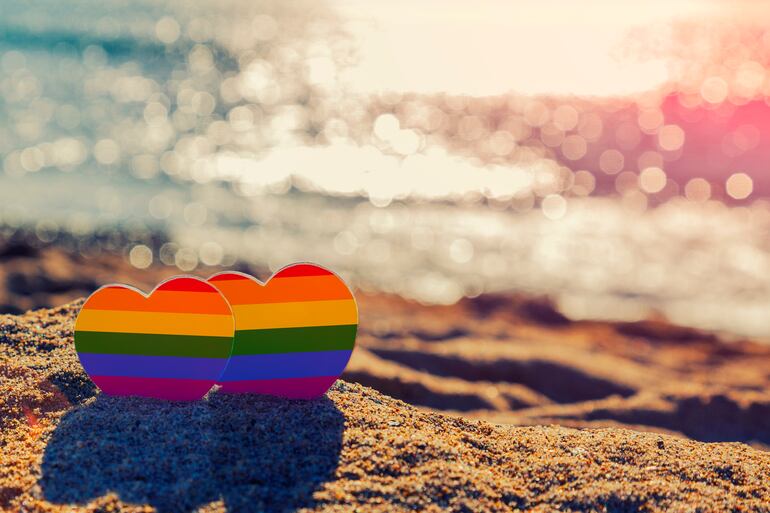 Dos corazones con los colores del arcoíris incrustados en la arena de una playa, frente a un curso de agua.