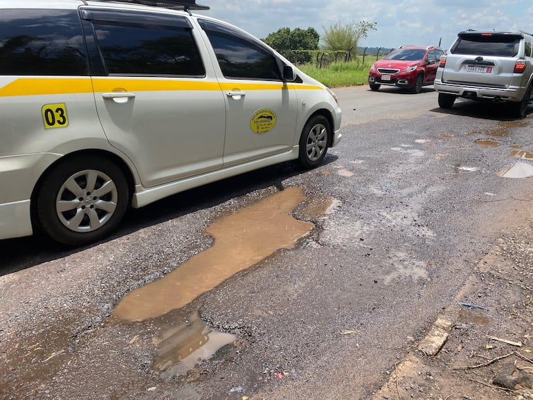 Vehículos, incluyendo un taxi de ruta, circulan con dificultad entre los baches llenos de agua sobre la deteriorada ruta departamental D027.