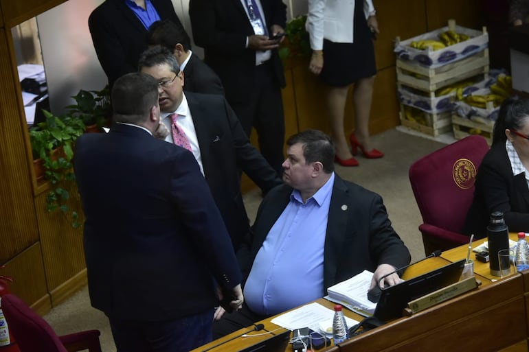 Un hombre obeso de prominente estómago, con camisa celeste y saco, está sentado frente a un escritorio sobre el que hay una computadora portátil. De pie junto a él hay dos hombres más parados hablando y en el fondo se ve parcialmente a otras tres personas y tres cajones de banana.