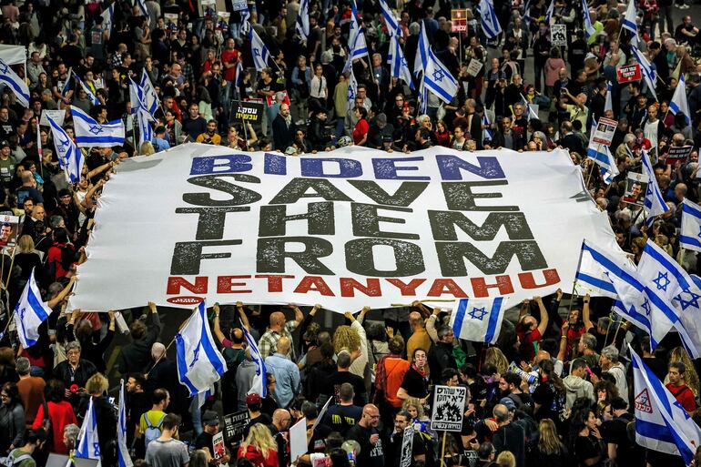 La gente levanta una gran pancarta pidiendo al presidente de los Estados Unidos que presione al gobierno israelí para que tome medidas para devolver a los rehenes mantenidos cautivos por militantes palestinos en la Franja de Gaza desde los ataques del 7 de octubre, durante una manifestación antigubernamental cerca de la sede del Ministerio de Defensa en Tel Aviv.