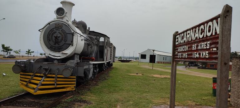 En primer plano el cartel indicador de la estación “Encarnación”, una antigua locomotora de vapor, y al fondo, frente a un galpón que representa el taller del ferrocarril, se encuentra la plataforma de giro.