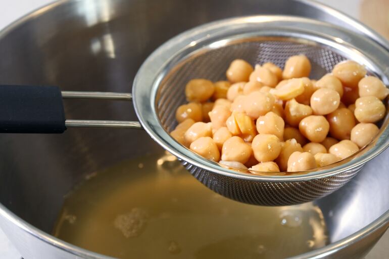 El contenido de una lata de garbanzos se pasa por un colador y se recoge el agua en un bol. Este líquido se llama aquafaba y es el ingrediente principal de las "claras" batidas veganas.