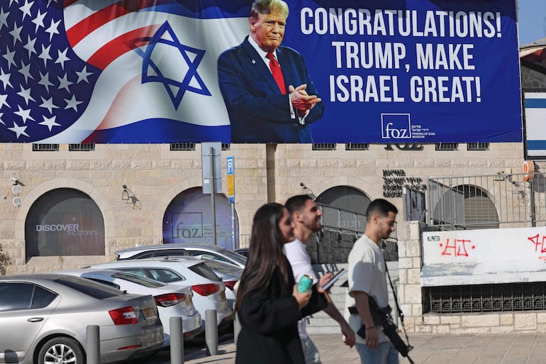 Tres personas, una de ellas armada, caminan frente a un cartel que celebra la victoria de Donald Trump en las elecciones estadounidenses, en Jerusalén.