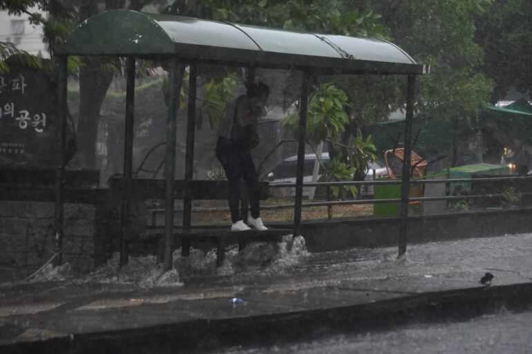 Una pasajera se resguarda precariamente de la lluvia parada en el banco de una parada de colectivos, mientras el raudal corre calle abajo.