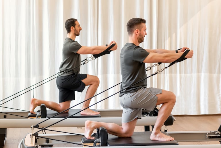 Dos hombres realizan ejercicios de estiramiento en un reformer de pilates.