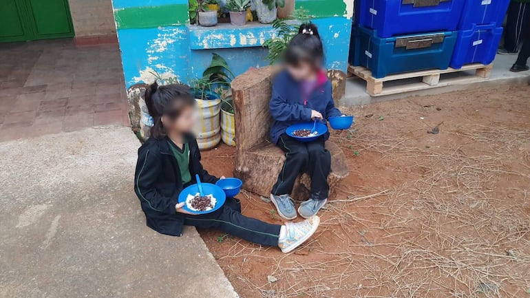 Una niña sentada en un tronco y otra, en el piso, durante el almuerzo escolar en Capiatá.