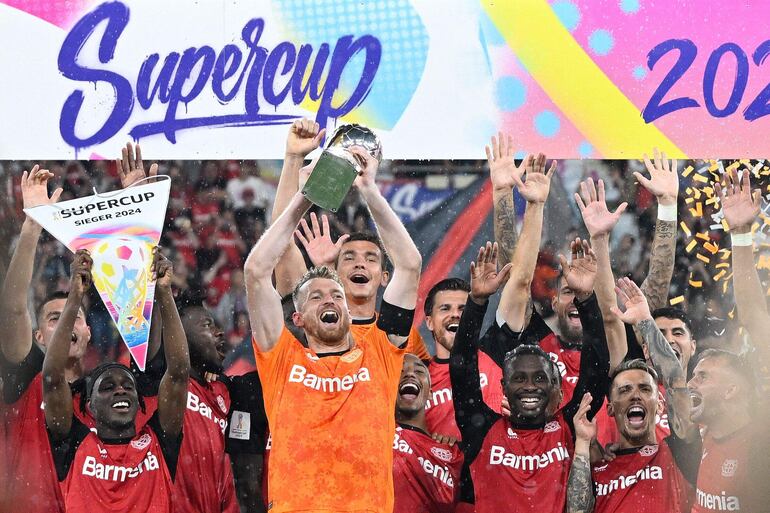 TOPSHOT - Bayer Leverkusen's Finnish goalkeeper #01 Lukas Hradecky and his teammates celebrate with the trophy after winning the penalty shoot-out of the German Supercup football match between Bayer 04 Leverkusen and VfB Stuttgart in Leverkusen, western Germany on August 17, 2024. (Photo by Sascha Schuermann / AFP) / DFL REGULATIONS PROHIBIT ANY USE OF PHOTOGRAPHS AS IMAGE SEQUENCES AND/OR QUASI-VIDEO