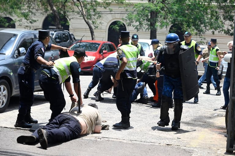 Con una cachiporra contra la espalda un policía inmoviliza a un funcionario de la ANDE en el suelo, mientras otro grupo de policías le da  patadas y golpes de cachiporra a otro manifestante.