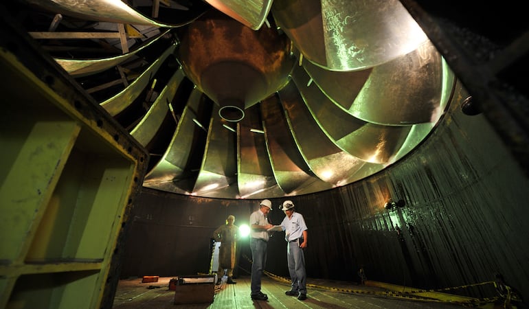 El tamaño de los técnicos y el de la pieza de una de las turbinas de la central hidroeléctrica.