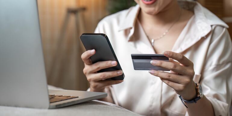 Una mujer con un teléfono celular en una mano y una tarjeta de crédito, frente a una laptop