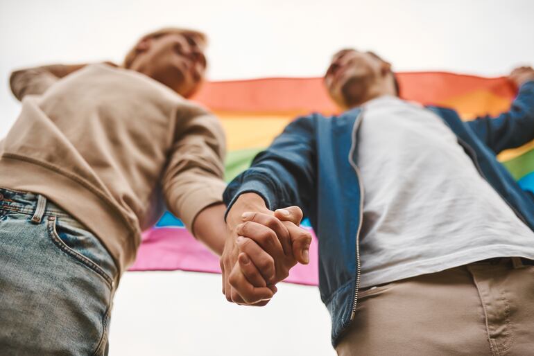 Imagen de referencia: dos personas agarrándose de la mano con una bandera del orgullo LGBTI de fondo.