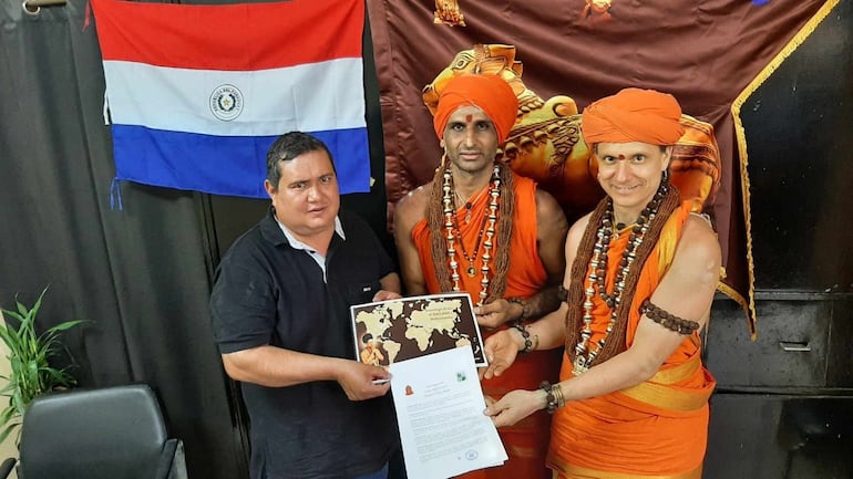 Un hombre moreno y petiso con sobrepeso sostiene un mapa y un documento. A su lado, dos hombres ataviados con telas de color anaranjado, turbantes, collares y brazaletes. De fondo, colgada la bandera de Paraguay, roja, blanca y azul y una bandera marrón con la figura de una diosa y un animal en dorado.