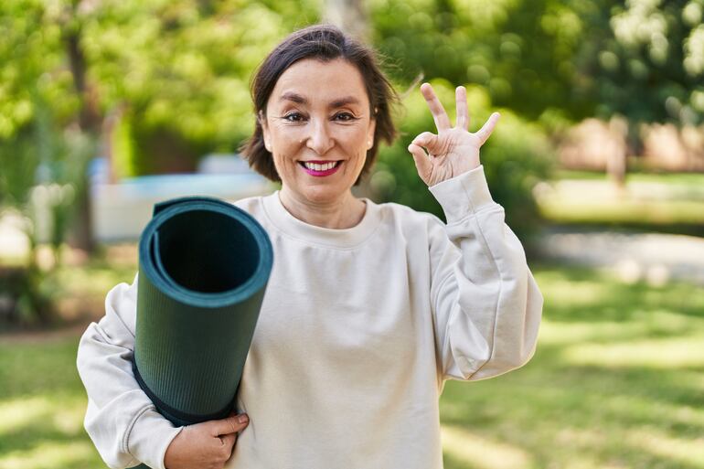 Una mujer de mediana edad sonrie con un mat de yoga enrrollado.