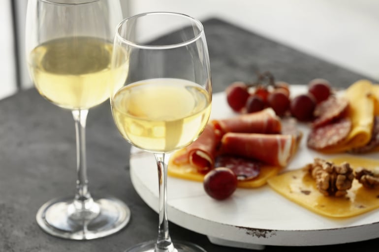 dos copas de vino blanco junto a una tabla quesos y encurtidos