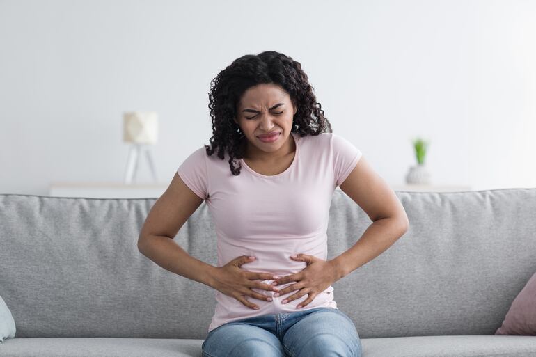 Una mujer morena se toca el estómago y frunce el seño por el dolor que siente.