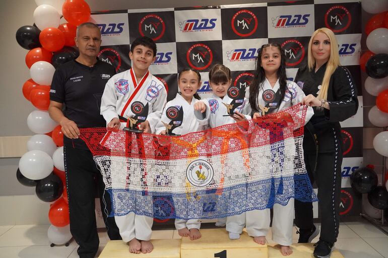 En la imagen de izquierda a derecha, el GM César Ozuna, Santiago Noé, Milena Recalde, Alethia Amarilla, Zoe Noé y la prof. Angelike Augsten posan con la bandera paraguaya en São Paulo.