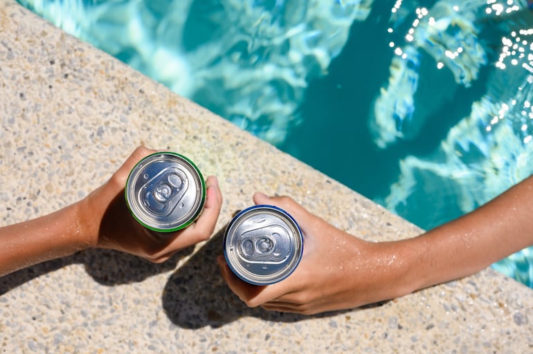 Dos manos de dos personas sosteniendo latas de cerveza en el borde de una pisicina.